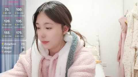 A video of an East Asian woman with long black hair, wearing a fluffy pink bathrobe, standing in a modern, light-colored bathroom.