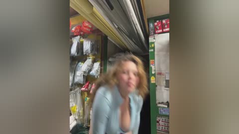 Video of a blonde woman in a low-cut, light blue shirt, blurred, standing in a cluttered, dimly lit pharmacy with shelves of products and a green wall.