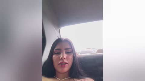 A video of a Latina woman with long dark hair, wearing a pink top and necklace, seated in a dimly lit car, eyes closed, lips slightly parted.