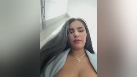 Video of a young woman with long, straight, dark hair, light skin, wearing a light blue blazer, unbuttoned, exposing cleavage. She has a neutral expression, standing in a dimly lit room with white walls and a window.