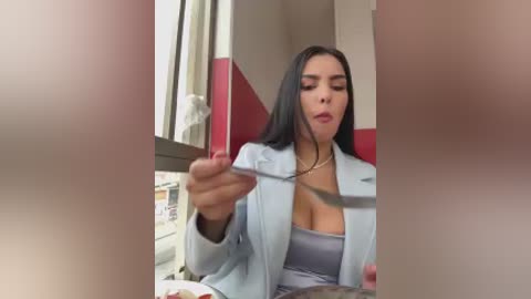 Video of a woman with long dark hair, wearing a light blue blazer over a low-cut gray top, seated in a red and white tiled bathroom, holding a cigarette.