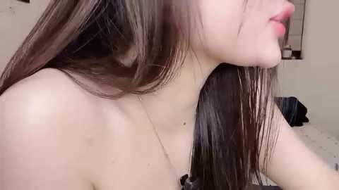 Video of a light-skinned woman with long, straight, dark brown hair, slightly parted lips, and bare shoulders, wearing a delicate necklace. Background shows a simple, indoor setting with beige walls and a blurred window.