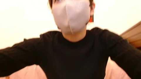 Video of a person wearing a white face mask, black long-sleeve shirt, and short brown hair, taken from a low angle, focusing on their upper body. The background is a blurred beige wall.