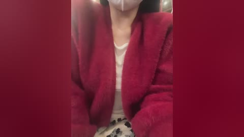 Video of a person in a red fleece jacket, white shirt, and black pants, wearing a white face mask, seated indoors, with blurred background.