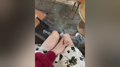 Video of a person with pale skin wearing a white dress with black floral print, sitting on a chair with red socks, surrounded by wooden chairs and a table in a cluttered room.