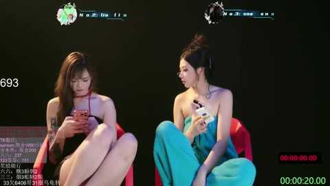 Video of two women with pale skin, one with long brown hair, the other with black hair in a bun, sitting on red chairs, holding phones, wearing black and turquoise outfits, against a dark background.