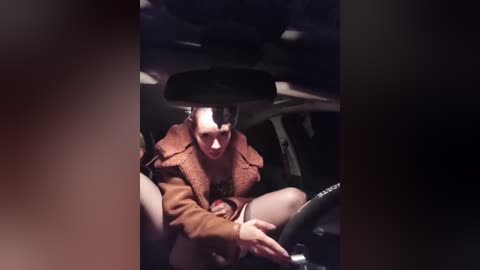 Video of a woman in a car, wearing a brown jacket, looking distressed, with a bandage on her head, sitting on the car's floor, hands resting on the steering wheel.