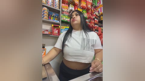 A video of a Latina woman with long black hair and medium skin tone, wearing a white crop top and black pants, standing behind a convenience store counter filled with colorful snacks.