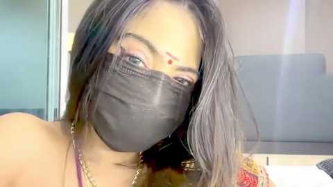 Media: A close-up video of a South Asian woman with long, dark hair, wearing a black face mask, red bindi, and gold chain necklace. Background shows a grey sofa and a partially visible TV.