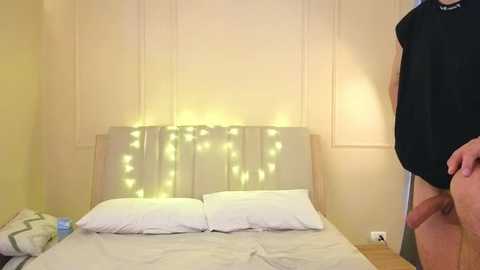 Video of a man standing beside a bed with white sheets and a \"DADDY\" sign, illuminated by string lights.
