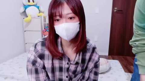 A video of an Asian woman with straight, shoulder-length red hair, wearing a white face mask, plaid shirt, and sitting on a bed in a modest, white-walled bedroom.