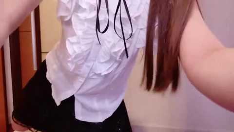 Video of a light-skinned woman with long brown hair, wearing a white ruffled blouse with black ribbons and a black mini skirt, standing indoors against a neutral background.