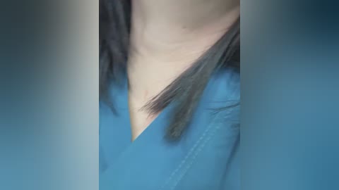 A close-up video of a person's neck and upper chest. The person has light skin and a black, straight hair extension draped over their shoulder. They wear a light blue denim shirt. The background is blurred and indistinct.
