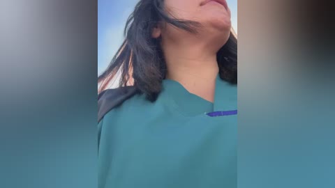 Video of a person with medium-length dark hair, wearing teal scrubs, looking up. Background is blurred, with a gradient of blue and beige colors. The image has a soft, dreamy aesthetic.