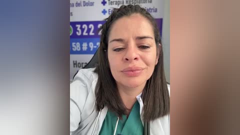Video of a middle-aged woman with straight, dark brown hair, wearing a white lab coat over a green shirt, standing in a hospital room with a blurred background displaying medical information.