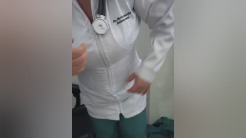Video of a woman in a white lab coat, with a stethoscope around her neck, holding her breasts. She wears teal scrubs. Background shows a blurred medical setting.