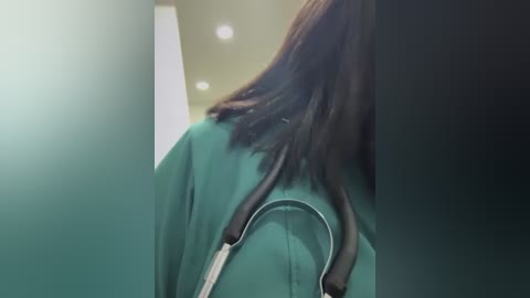 A video capturing a medical professional's back, wearing a teal hospital gown with a stethoscope draped around the neck. The background is a sterile, white-tiled hallway with recessed lights, conveying a clinical setting.