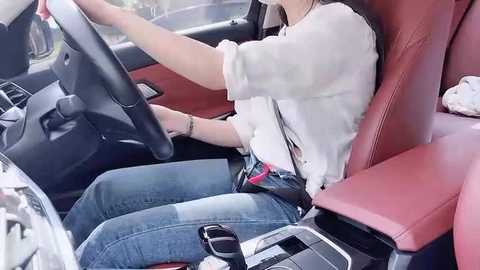 Video of a young woman with light skin, wearing a white shirt and blue jeans, driving a car with a red interior, seen from the passenger side, holding a pink cellphone.