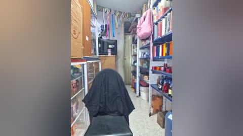 Video of a small, dimly lit storage room with a person in a black robe standing among shelves of household items, clothes hanging from the ceiling, and a white toilet in the background.