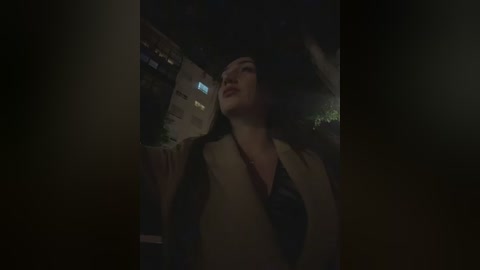 A dimly lit video shows a woman with long black hair, wearing a beige blazer and a black shirt, looking upward with a serene expression. The background is dark and indistinct, suggesting an urban night scene.