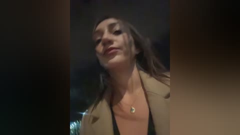 Video of a young woman with light skin, long dark hair, wearing a tan blazer and black top, standing outdoors at night with blurred, dark background.