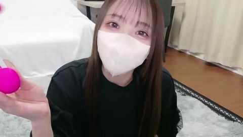 Video of an Asian woman with long brown hair, wearing a black top and white face mask, kneeling on a light grey carpet in a room with a bed and wooden floor.