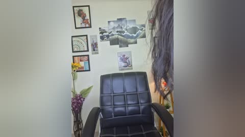 A video of a cozy, minimalist room featuring a black leather recliner, framed artwork, a floral arrangement, and a vibrant tapestry on a white wall.