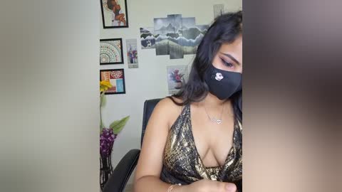 Video of a woman with long black hair, wearing a black face mask, a revealing snakeskin-patterned halter top, and a delicate necklace, seated at a desk in a colorful, eclectic room with art and floral decorations.