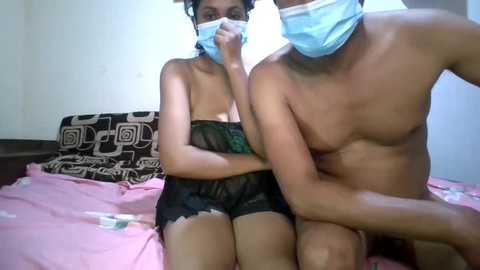 Video of a topless, mask-wearing couple on a pink bed, with a black-patterned pillow. The woman, wearing a sheer black dress, has a slender physique, and the man, shirtless, has a muscular build.