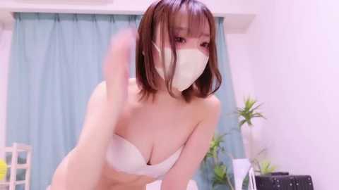A video of an East Asian woman with shoulder-length brown hair, wearing a white bra and face mask, bending forward in a room with light blue curtains, potted plants, and a white chair.
