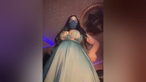 Video of a curvy woman with long black hair, wearing a teal gown and blue face mask, standing in a dimly lit room with a brick ceiling and circular fan.