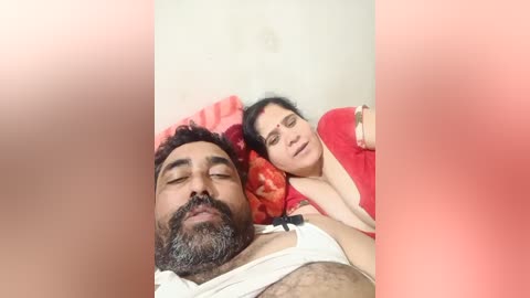 A video of a bearded man with dark skin, wearing a white shirt, and a woman with fair skin, wearing a red sari, sleeping side by side on a bed with pink and red pillows.