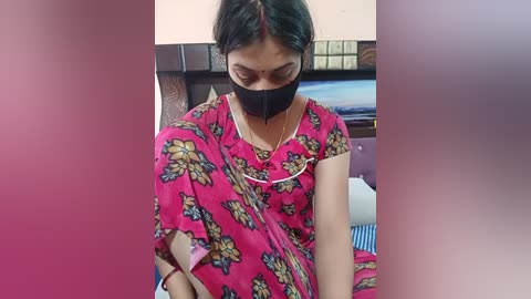 Video of a South Asian woman with medium skin tone, wearing a pink sari with floral design, black face mask, and a gold chain, seated indoors.
