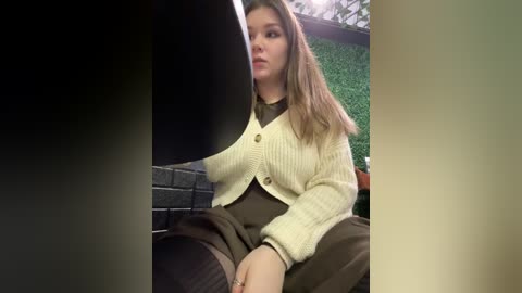 Video of a young Caucasian woman with long, straight light brown hair, wearing a cream knitted cardigan over a dark blouse, sitting on a dark couch. Background includes green faux grass and a brick wall.