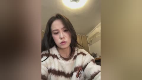 Video of a woman with long black hair, wearing a white and brown striped sweater, sitting in a dimly lit room with beige walls and a ceiling light.