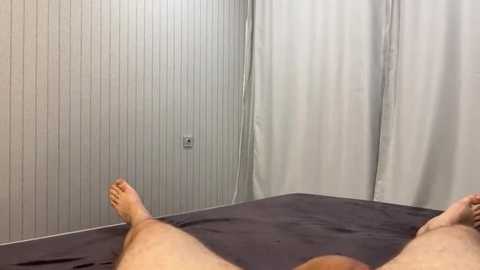 Video of a person lying on a bed with gray sheets, legs stretched out. Background features white paneled walls and a white curtain.