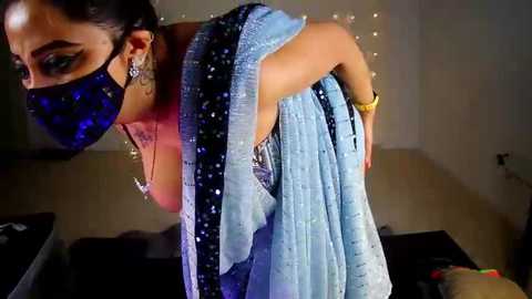 Video of a woman with dark skin, wearing a blue saree, black face mask, and glittery earrings, in a dimly lit room with soft lighting and a dark background.
