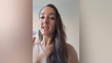 Video of a topless, fair-skinned woman with long, straight brown hair and a slender physique, standing in a dimly lit room with blurred, beige walls.