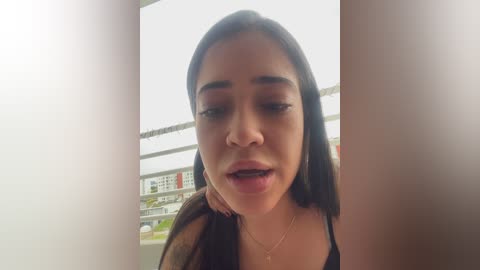 A video of a young woman with medium-brown skin and long dark hair, wearing a black top and gold necklace, smiling outdoors near a modern building.