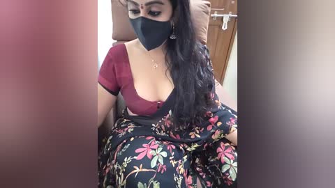 Video of a South Asian woman with long black hair, wearing a floral black saree, black mask, and maroon top, seated indoors on a beige couch.