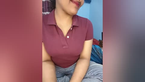 Video of a woman with light brown skin and dark hair, wearing a burgundy polo shirt and gray shorts, sitting on a bed with blue and red bedding in the background.