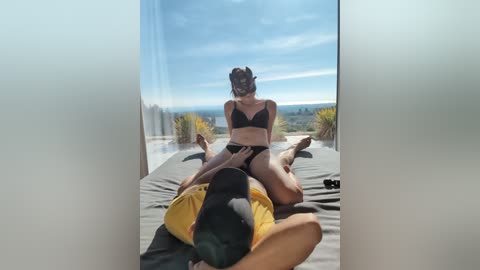 A video captures a woman in a black bra and panties, sitting on a man lying on a bed. They are in a modern room with a large window displaying a scenic landscape.