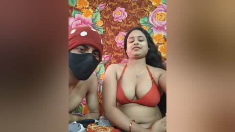 Video of two South Asian women, one with a red headscarf and mask, the other in a red bikini, lying on a colorful floral bedspread, background blurred.