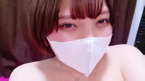 Video of an Asian woman with short brown hair, wearing a white face mask, and a pink top. She has light skin, dark eyes, and is looking directly at the camera.