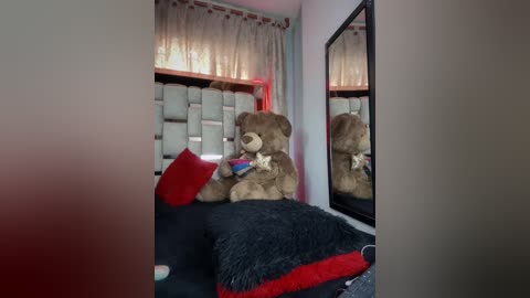 Video of a cozy, dimly lit bedroom with a large teddy bear on a black bed with red and blue pillows, a tufted headboard, and a mirror reflecting the teddy bear and bed.