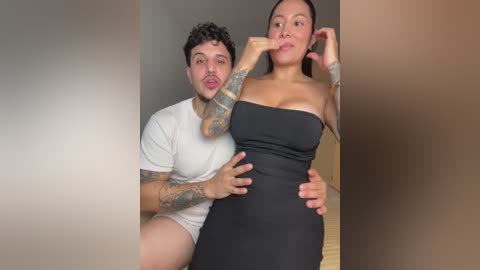 Video of a tattooed man in white shorts and a woman in a black dress, both posing intimately indoors.