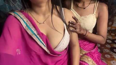 A video of two South Asian women, one in a pink sari with a white bra visible, the other in a beige lace top, both with dark skin, sitting closely.