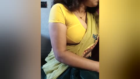 A video of a woman with medium brown skin, wearing a yellow shirt that reveals cleavage, sitting on a dark couch. Her long dark hair is partly visible.