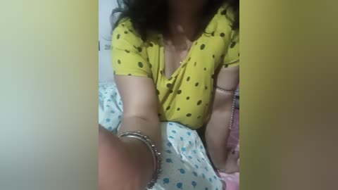 Video of a woman with medium skin tone, wearing a yellow polka-dotted blouse and bracelets, lying on a bed with blue and white polka-dotted sheets, partially obscured by a yellow gradient overlay.