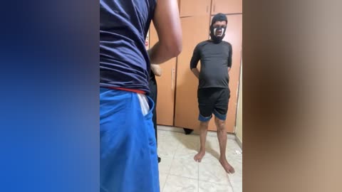 Video of a barefoot man in a black T-shirt and blue shorts standing in a tiled room with wooden cabinets and a beige floor.